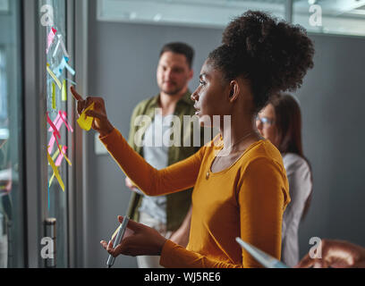 Vista laterale di un giovane africano imprenditrice controllo e blocco note su vetro alla riunione in ufficio moderno Foto Stock