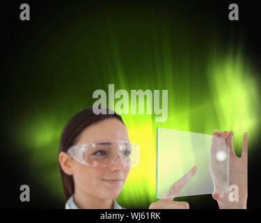 Immagine composita di bruna medico in occhiali protettivi tenendo premuto lo schermo virtuale su sfondo nero con incandescente verde all'interno Foto Stock