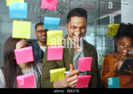 Gruppo di sorridere casual la gente di affari di pianificazione e di progetto di scrittura e idee su foglietti adesivi incollato sulla parete in vetro presso un ufficio moderno Foto Stock