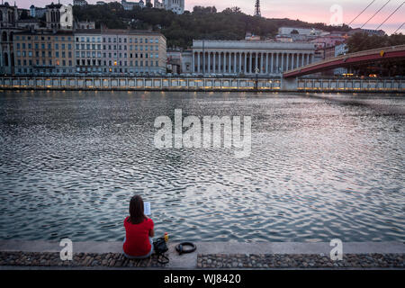 Lione, Francia - Luglio 17, 2019: giovane donna leggendo un libro e rilassante sulla sponda del fiume Saone a Lione, al tramonto, con colline de Fourviere hill b Foto Stock