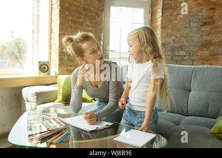 Femmina insegnante caucasica e bambina, o la mamma e figlia. Homeschooling. Seduti sul divano e disegno con vernici, scrittura, conversazione e divertirsi. Istruzione, scuola, studiando concetto. Foto Stock