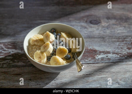 Una ciotola di porridge di avena guarnita con fette di banana e mirtilli. Il vaso è su un rustico sfondo legno consentendo l'abbondanza di spazio copia. Foto Stock