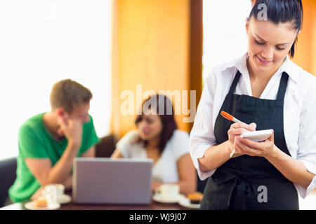 Bella cameriera di scrittura di un ordine con due studenti che usano il computer portatile presso la caffetteria Foto Stock