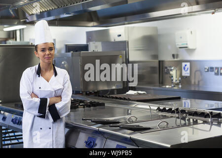 Giovani chef felice in piedi accanto alla superficie di lavoro con le braccia incrociate in cucina professionale Foto Stock