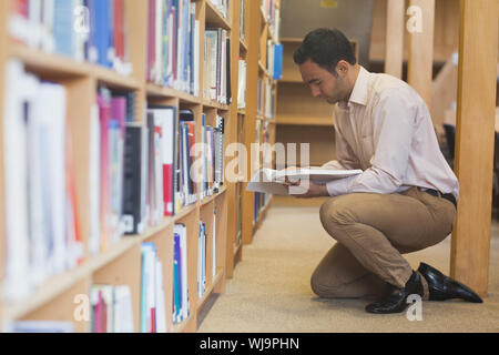 Attraente uomo informale la lettura di un libro in biblioteca in ginocchio davanti agli scaffali Foto Stock