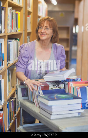 Femmina bibliotecario maturo restituendo libri in biblioteca sorridente in telecamera Foto Stock