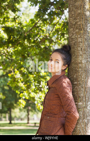 Vista laterale di attraenti brunette donna appoggiata contro un albero sorridente in telecamera Foto Stock