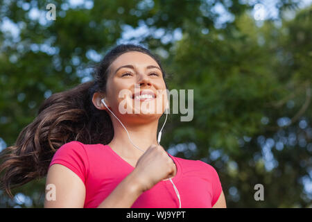 Gioioso donna sportiva jogging in un parco e ascolto di musica Foto Stock