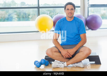 A piena lunghezza Ritratto di un giovane uomo seduto con manubri in studio fitness Foto Stock