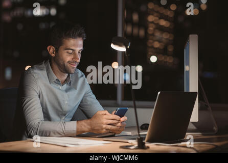 Sorridente giovane imprenditore la lettura di un messaggio di testo mentre si lavora fino a tardi Foto Stock