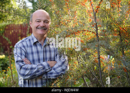 Senior ispanico uomo in piedi nel suo giardino vicino al mare frangola sorridente. Foto Stock