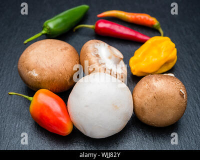 Crimini di funghi in marrone e bianco e peperoncino piccante di diversi colori su nero ardesia. Messa a fuoco selettiva in primo piano. Foto Stock