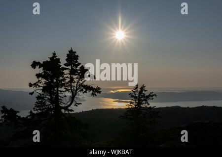 Tramonto su i fiordi al di fuori di Bergen, Norvegia, incorniciato tra alberi sempreverdi in silhouette con il sole guardando come una raggiera. Foto Stock