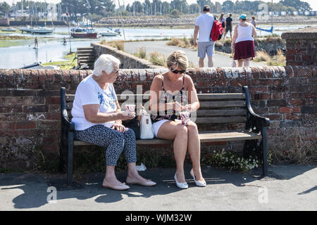 Una donna di mezza età e una signora anziana seduta su una panchina nel parco a bere un caffè da asporto Foto Stock