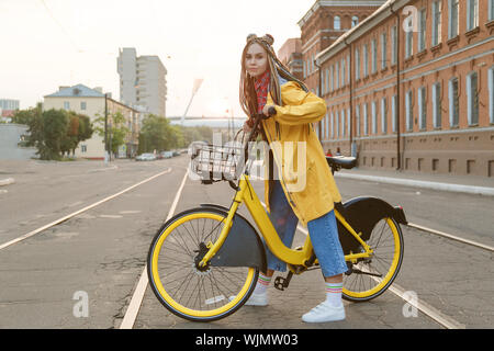 Giovane donna indossa cappotto giallo e colorato pigtail, equitazione bicicletta in città. Foto Stock