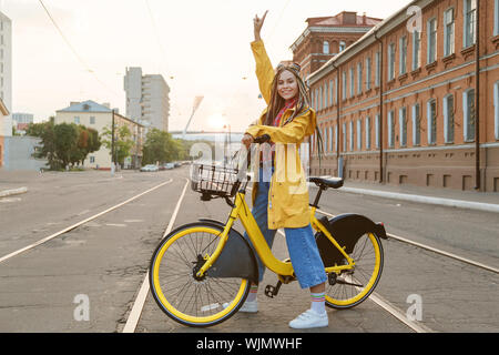 Giovane donna indossa cappotto giallo e colorato pigtail, equitazione bicicletta in città. Foto Stock
