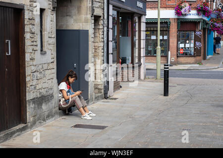 Una donna asiatica nel suo ventenne guardando il suo telefono cellulare mentre seduto su un gradino al di fuori di un negozio su un high street Foto Stock