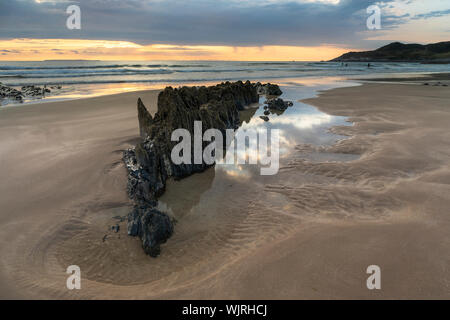 Formazione di roccia a Combesgate Beach in North Devon circondato da acqua che riflette il cielo con la bassa marea con surfers in distanza. Foto Stock