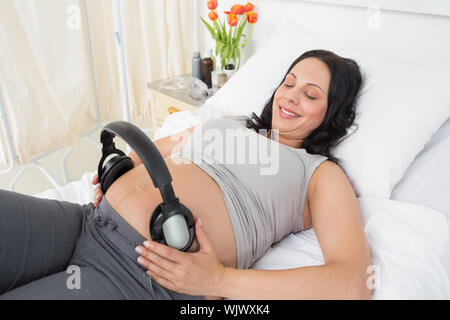 Sorridente donna in attesa di mettere le cuffie sul suo ventre mentre giaceva a letto di ospedale Foto Stock
