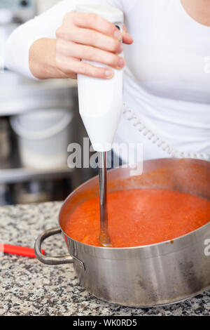 Lo chef donna sbattere bollito di pomodoro in una pentola per una loro riduzione in una purea di base per zuppe o salse con un dispositivo azionato a batteria frusta Foto Stock