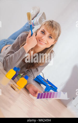 Bella donna sorridente giace sul pavimento in legno tenendo il pennello e la tavolozza dei colori Foto Stock