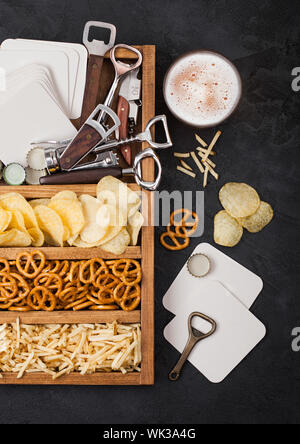 Bicchiere di craft birra lager e apriporta con scatola di spuntini sulla cucina nera sfondo tabella. Pretzel e patatine e salato bastoncini di patata in woode vintage Foto Stock