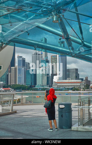 Un giovane turista femminile coperto con shiny destro rosso scialle di Marina Bay, Singapore, fotografando la skyline della citta'; sopra: parte del ponte di Helix Foto Stock