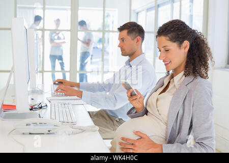 Casual incinta imprenditrice texting sul telefono alla scrivania in ufficio Foto Stock