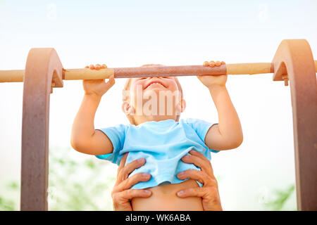 Ragazzino con padri aiutare a recuperare il ritardo accumulato sulla barra orizzontale, attiva l'infanzia, carino piccolo acrobat, allenamento sul cortile, summer camp concept Foto Stock