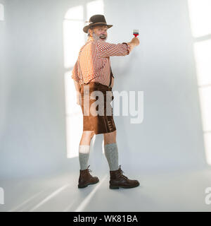 Ritratto di Oktoberfest senior uomo in hat, indossando i tradizionali abiti bavarese. Maschio a tutta lunghezza shot presso lo studio su sfondo bianco. La celebrazione, vacanze, festival concetto. Verniciatura di un muro. Foto Stock