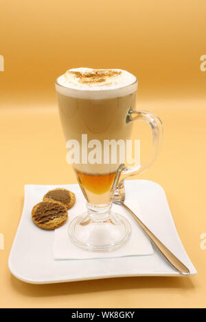 Tazza in vetro di latte macchiato a strati con schiuma di latte e cannella, servita con biscotti e cucchiaio su un piatto bianco, isolato su sfondo arancione Foto Stock