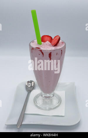 Bicchiere alto di frullato di fragole con fette di fragola fresche e paglia verde, servito su un piatto bianco con un cucchiaio Foto Stock