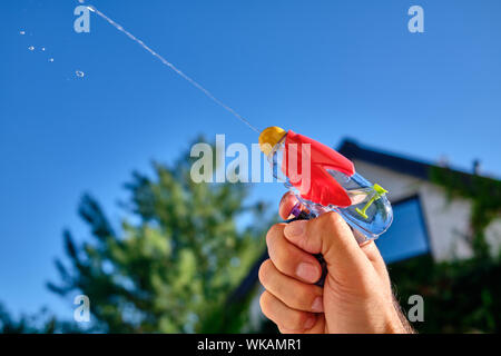 Acqua colorata pistola spruzzatura fuori un getto di acqua in un maschio caucasico mano nella parte anteriore di una casa e il cielo blu di una soleggiata giornata estiva Foto Stock