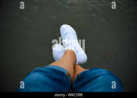 Gambe in jeans sneakers bianco sul fiume vista dall'alto. Foto Stock