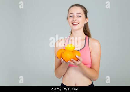 Giovane donna in usura fitness contiene frutta arance e limone nelle sue mani, isolata su uno sfondo grigio. Foto Stock