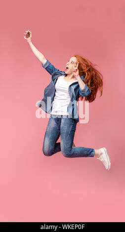 Felice redhead girl jumping e rendendo selfie sullo smartphone Foto Stock