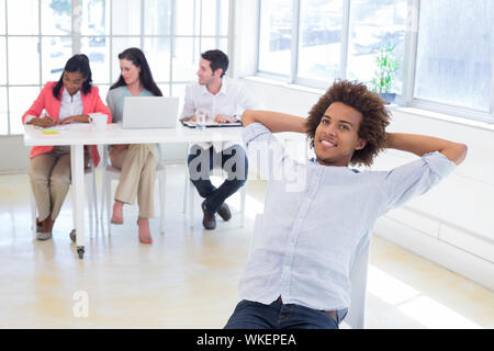 Imprenditore relax su una sedia girevole a sorridere mentre i suoi colleghi stanno lavorando dietro di lui Foto Stock