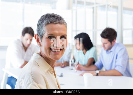 Attraente imprenditrice sorridente verso la telecamera sul posto di lavoro con altri colleghi Foto Stock