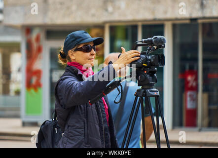 Wolfsburg, Germania, Settembre 1, 2019: giovane camerawoman in blu giacca con cappuccio e occhiali da sole per la configurazione del suo videocamera montata su di un cavalletto Foto Stock