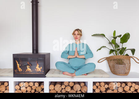 Ragazza incinta in blu sportswear si siede nella posizione del loto fare yoga in interni accoglienti Foto Stock