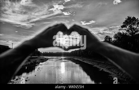 Ricordi estivi concetto. Primi posti per romantiche data. La luce del sole al tramonto atmosfera romantica. Mani maschio a forma di cuore simbolo del gesto di amore e romanticismo. Cuore gesto davanti al tramonto sopra il fiume. Foto Stock