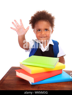 Felice preschooler africani seduti dietro una scrivania con molti libri su di esso, isolato su sfondo bianco, sollevata la mano e dire ciao, torna a scuola concep Foto Stock