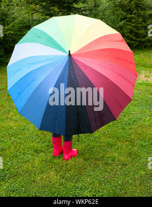 Bambina rosso da indossare stivali di gomma di nascondersi dietro ombrello coloratissimo sotto la pioggia Foto Stock