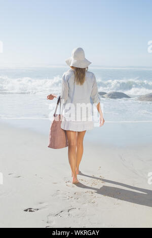 Donna bionda nel cappello che trasportano borsa da spiaggia che si affaccia sul mare da una giornata di sole Foto Stock