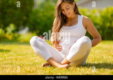 Donna incinta in bianco seduto sull'erba all'esterno tenendo la pancia Foto Stock