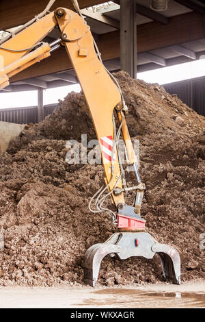 Per impieghi pesanti escavatore facendo movimento terra undercover parcheggiata in un magazzino aperto davanti a un enorme mucchio di terra bruna per la costruzione Foto Stock