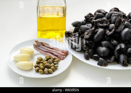 Ingredienti per la tapenade: olive nere snocciolate sulla piastra bianca, olio di oliva in una bottiglia di vetro, tre spicchi dâ aglio, alcuni filetti di acciughe e capperi. Foto Stock