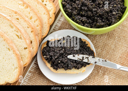 Sandwich tapenade e coltello da tavola su un sauser, verde ciotola con tapenade fresco e fette di pane di grano su una tela Tovagliolo da tavola. Cuocere a casa. Foto Stock