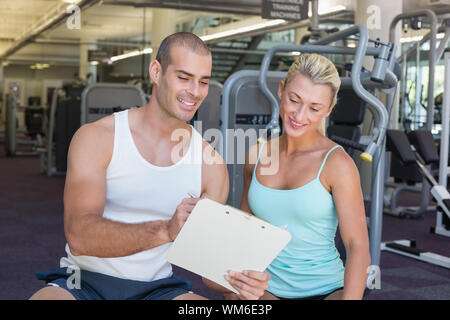 Bella giovane donna a discutere la sua performance negli appunti con un trainer in palestra Foto Stock