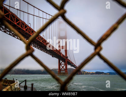 Il famoso Golden Gate bridge visto attraverso un arrugginito catena collegamento recinto su un nuvoloso giorno di estate con bassa appendere la laminazione di nebbia a San Francisco, California Foto Stock